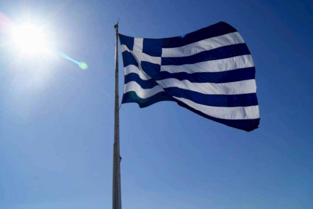 Greece's national flag waving against a clear blue sky and bright sun.